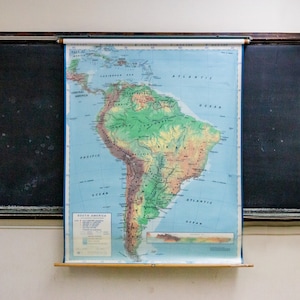 May include: A vintage classroom map of South America with a blue background and green, brown, and yellow landmasses. The map is mounted on a wooden roller and is hanging on a black chalkboard.