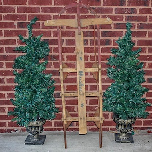 May include: A vintage wooden sled with red paint and the word "Huddleston" printed on the side. The sled is flanked by two small artificial Christmas trees in pots.