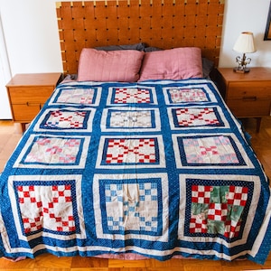 May include: A blue and white patchwork quilt with a red, white, and blue checkered pattern. The quilt is made of squares and is laid out on a bed with a wooden headboard.