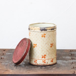 May include: A vintage, white metal canister with a red lid and a floral pattern in orange. The canister has a white background with orange dots and orange floral designs.