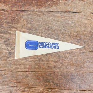 May include: A cream-coloured pennant featuring the words "Vancouver Canucks" in blue, and a blue hockey stick and puck logo. The triangular fabric pennant is set against a wooden background.