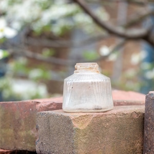May include: A clear glass jar with a textured surface sits on top of a stack of red bricks. The jar is empty and has a slightly chipped rim.