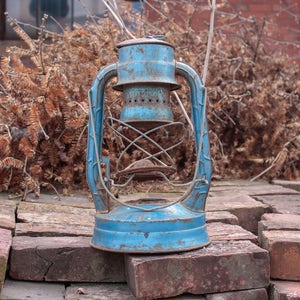 May include: A blue metal kerosene lantern with a wire cage around the glass. The lantern is sitting on a pile of red bricks.