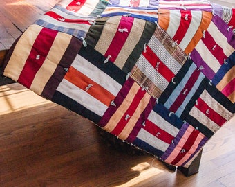May include: A patchwork quilt made from various fabrics in shades of red, blue, white, and brown. The quilt is made of squares and rectangles, and the seams are visible. The quilt is folded and draped over a wooden table.