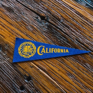 May include: A blue pennant with gold lettering and a university seal. The pennant reads "CALIFORNIA" and features the seal of the University of California. The pennant is on a wooden surface.