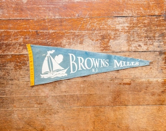 May include: A vintage pennant in a triangular shape, featuring a sailboat illustration and the text "BROWNS MILLS N.J." in white on a light blue background. The pennant has a yellow border and rests on a wooden surface.