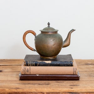 May include: A copper teapot with a wooden handle sits on top of a stack of three books. The books are on a wooden table.