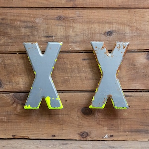 May include: Two large, silver metal letters spelling "XX" are attached to a wooden wall. The letters have a worn, distressed look and are painted with yellow accents.
