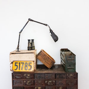May include: A vintage metal industrial style drawer unit with a yellow license plate reading "51785" on top.  The drawer unit is topped with a wooden crate, a smaller metal crate, and a black metal lamp with a black shade.  There are also binoculars on top of the wooden crate.