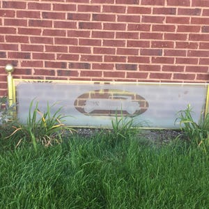 May include: A gold-colored metal frame with a frosted glass panel. The glass panel has a silhouette of a car etched into it. The frame is mounted on a brick wall and is surrounded by green grass.