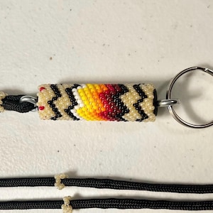 May include: Handmade beaded keychain with a cylindrical design. The central beadwork features a cream-colored base with black zigzag patterns and a gradient of yellow, orange, and red. A silver keyring is attached.
