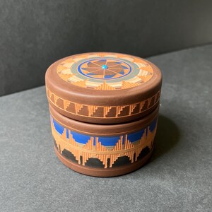 May include: A small, round, brown ceramic box with a lid. The box is decorated with intricate geometric patterns in shades of brown, blue, and turquoise. The lid features a central turquoise stone.
