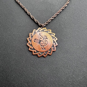 May include: Copper-toned pendant necklace featuring a stylized bird within a sunburst design. The pendant hangs from a matching rope chain. The necklace is displayed on a dark surface, highlighting the warm tones of the metal.