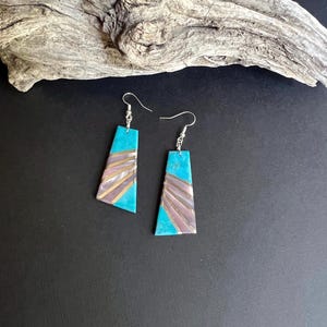 May include: A pair of turquoise and silver earrings with a geometric design. The earrings have a rectangular shape with a turquoise base and a layered design in shades of silver and purple. The earrings are suspended from silver hooks.