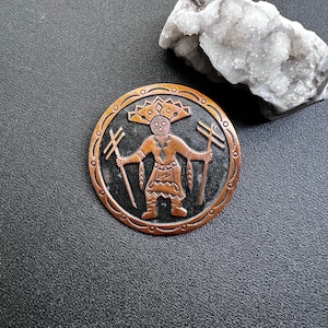 May include: A round copper pin with a Native American design. The pin features a stylized figure with a headdress and two staffs. The figure is surrounded by a circle of decorative elements.