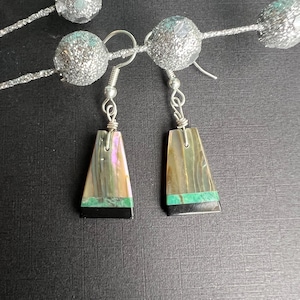 May include: A pair of handcrafted earrings with iridescent abalone shells. The earrings have a trapezoid shape, with a black base and a green accent. The silver-toned ear wires are visible. The background includes silver glittered spheres.