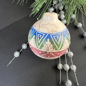 May include: A ceramic Christmas ornament with a cream-colored top and a patterned design in blue, green, and pink. The ornament has a crackled texture and is suspended by a metal loop. Silver beaded garland and pine branches are in the background.