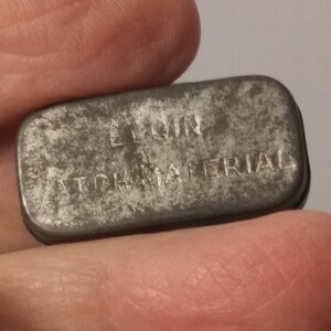 May include: A small, rectangular, metal tin with the words "ELGIN WATCH MATERIAL" embossed on the top.