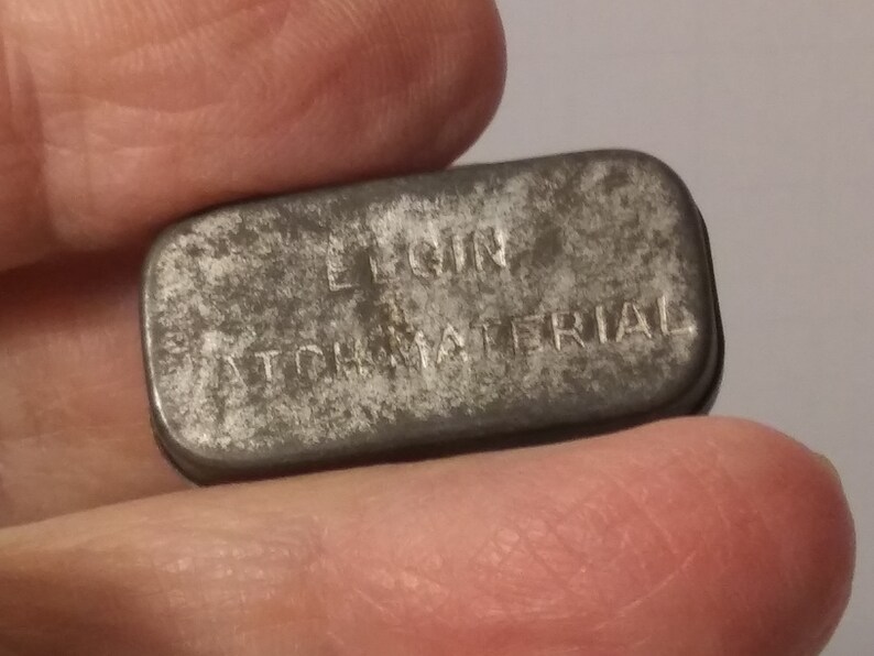 May include: A small, rectangular, metal tin with the words "ELGIN WATCH MATERIAL" embossed on the top.