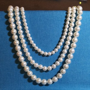 May include: A three-strand pearl necklace with white pearls of varying sizes. The necklace is displayed on a blue fabric background.