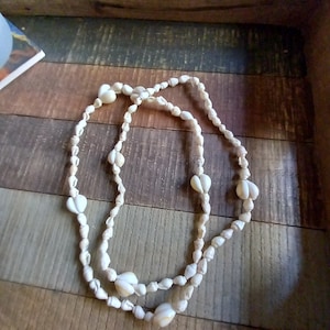 May include: A white shell necklace with a double strand of small shells and larger shells in the center. The necklace is on a wooden surface.