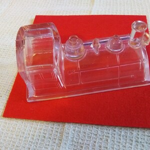 May include: A clear glass train engine figurine with a detailed design. The engine has a large round boiler and a tall chimney. The figurine is sitting on a red felt surface.