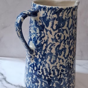 May include: A tall, ceramic pitcher with a large handle. The pitcher features a blue and white speckled pattern. The handle is curved and attached to the side of the pitcher. The pitcher has a spout for easy pouring.