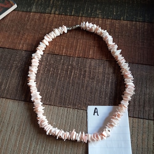 May include: A pink shell necklace with a silver clasp. The necklace is on a wooden surface with a white piece of paper with the letter "A" written on it.
