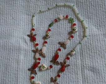 May include: A white, red, and green beaded necklace with angel wing charms and heart-shaped beads. The necklace is on a white textured surface.