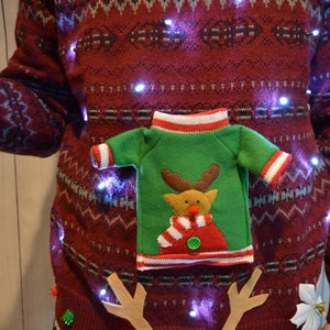May include: A festive burgundy Christmas sweater with a repeating white and blue pattern. The sweater features a green felt applique of a reindeer wearing a scarf and a miniature sweater. Small white lights are strung across the sweater.