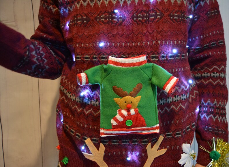 May include: A festive burgundy Christmas sweater with a repeating white and blue pattern. The sweater features a green felt applique of a reindeer wearing a scarf and a miniature sweater. Small white lights are strung across the sweater.