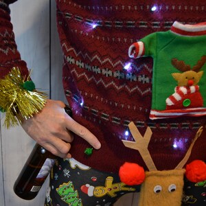 May include: A festive red Christmas sweater with a reindeer design and small lights. The sweater has a green pocket with a reindeer applique. The person is wearing gold tinsel wristbands and Christmas-themed pajama pants, holding a beer bottle.