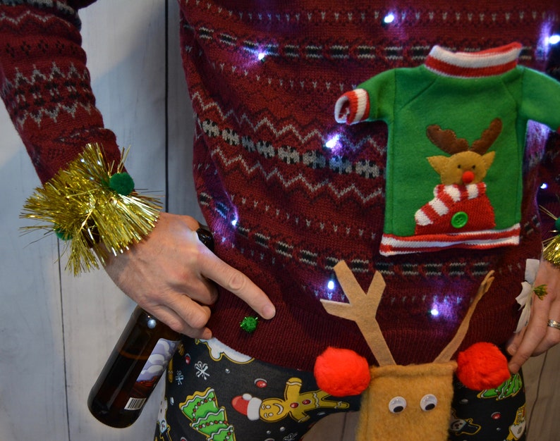 May include: A festive red Christmas sweater with a reindeer design and small lights. The sweater has a green pocket with a reindeer applique. The person is wearing gold tinsel wristbands and Christmas-themed pajama pants, holding a beer bottle.