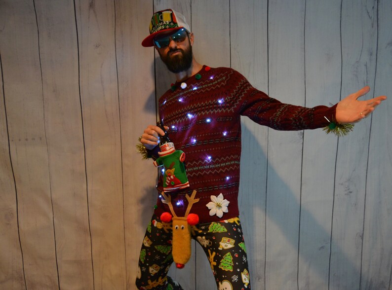 May include: A festive Christmas sweater with embedded lights, paired with patterned pants. The sweater features a reindeer-themed accessory and a decorative bottle holder. The outfit is completed with a cap and sunglasses, creating a humorous holiday ensemble.