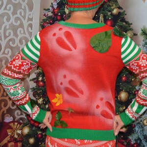 Puede incluir: Suéter rojo de Navidad con un patrón de copo de nieve blanco, ribetes verdes y huellas de reno blancas. El suéter tiene una hoja verde y una flor amarilla adheridas.