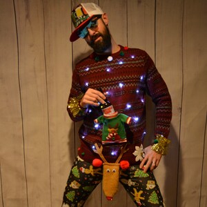 May include: A festive burgundy Christmas sweater with embedded lights, paired with patterned pants featuring Christmas trees and reindeer. The sweater has a beer bottle holder and a reindeer-themed accessory.