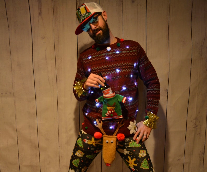 May include: A festive burgundy Christmas sweater with embedded lights, paired with patterned pants featuring Christmas trees and reindeer. The sweater has a beer bottle holder and a reindeer-themed accessory.
