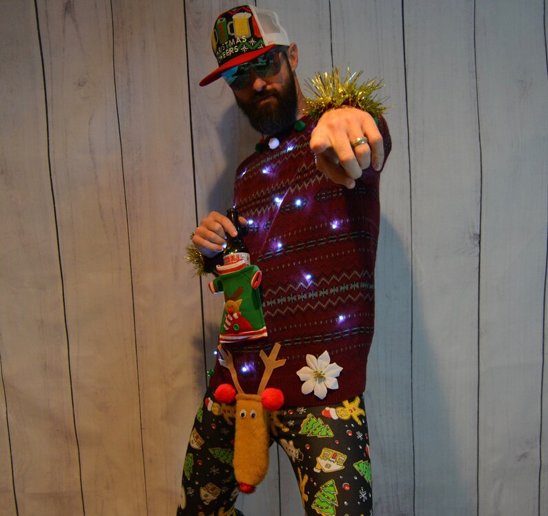 May include: A festive Christmas sweater with embedded lights, paired with patterned pants featuring Christmas trees and campers. The sweater is adorned with a white flower and a beer bottle cozy. A hat with the text "Christmas Beers" is worn.