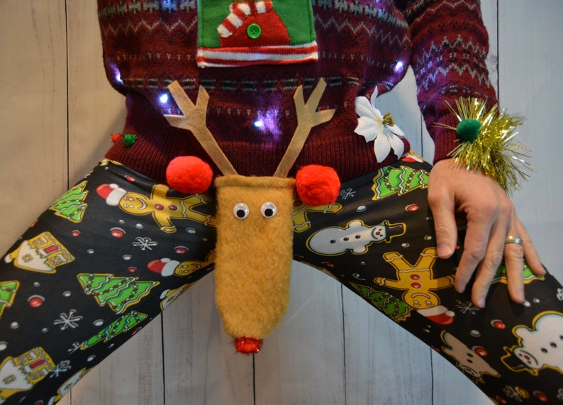 May include: A festive Christmas sweater with a reindeer-themed crotch decoration. The burgundy sweater has a green and white design, and is paired with black leggings patterned with gingerbread men, Christmas trees, and snowmen. The reindeer has googly eyes and red pom-pom antlers.