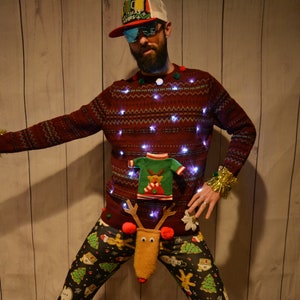May include: A festive Christmas sweater with embedded lights, a small sweater applique, and reindeer pants. The sweater is burgundy with a pattern, and the pants have a holiday print. The person is holding a beer bottle.