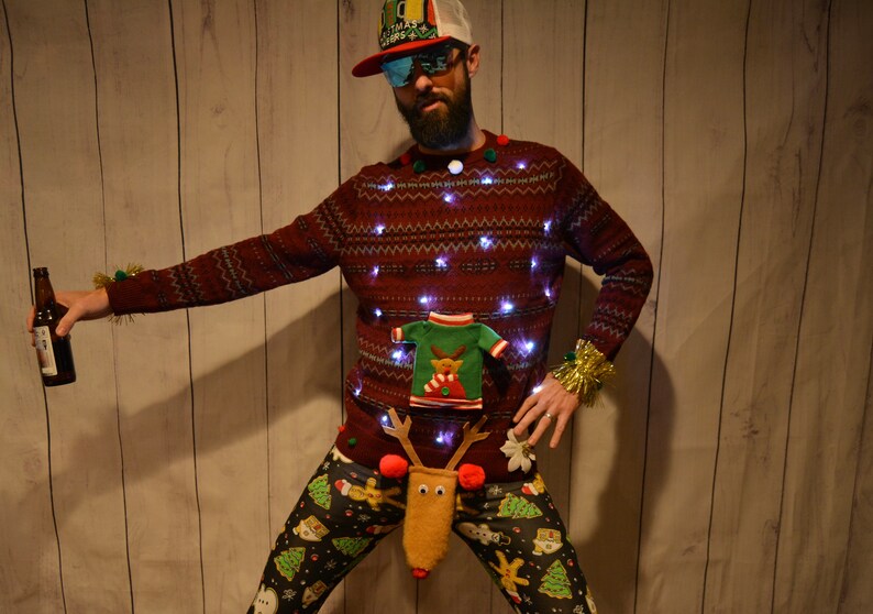 May include: A festive Christmas sweater with embedded lights, a small sweater applique, and reindeer pants. The sweater is burgundy with a pattern, and the pants have a holiday print. The person is holding a beer bottle.
