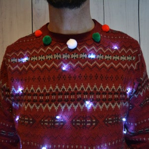 May include: A festive red sweater with a fair isle pattern and small white lights. The sweater is adorned with red, green, and white pom-poms around the neckline, creating a cheerful holiday look. The lights are evenly spaced across the front of the sweater.