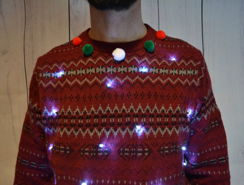 May include: A festive red sweater with a fair isle pattern and small white lights. The sweater is adorned with red, green, and white pom-poms around the neckline, creating a cheerful holiday look. The lights are evenly spaced across the front of the sweater.