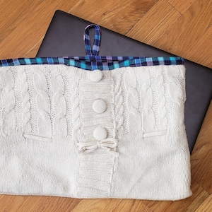 May include: White knitted laptop sleeve with a plaid fabric lining and a button closure. The sleeve is designed to protect a laptop from scratches and bumps.
