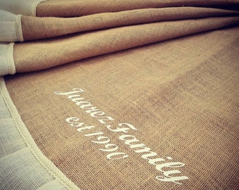 May include: A tan burlap table runner with white trim and the text "Juarez Family est 1990" in white script. The runner is folded, showing the texture of the burlap and the decorative trim.