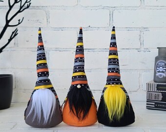 May include: Three Halloween gnomes with striped hats. The gnome on the left has a white beard and a brown body. The gnome in the middle has a black beard and an orange body. The gnome on the right has a yellow beard and a black body.