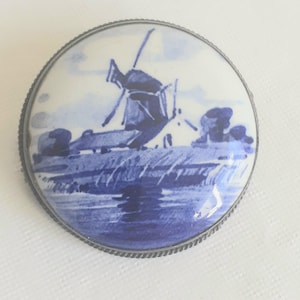 Puede incluir: Un broche vintage de porcelana azul y blanca con una escena de molino de viento. El broche está engastado en un marco de metal plateado.