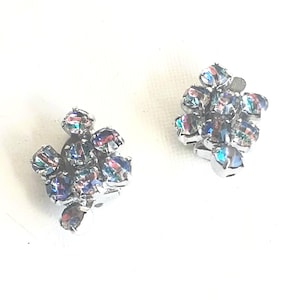 Puede incluir: Un par de pendientes vintage de clip. Cada pendiente presenta un grupo de piedras iridiscentes y facetadas engastadas en metal plateado. Las piedras reflejan colores como el azul, el morado y el verde, creando un efecto brillante. Los pendientes son para orejas no perforadas.