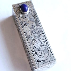 Könnte beinhalten: Ein rechteckiges Silberetui mit detaillierten floralen Gravuren. Auf der Oberseite ist ein kleiner, ovaler, tiefblauer Edelstein eingesetzt. Das Etui hat einen Klappdeckel und scheint aus Metall zu sein. Das Design ist detailliert und verziert.