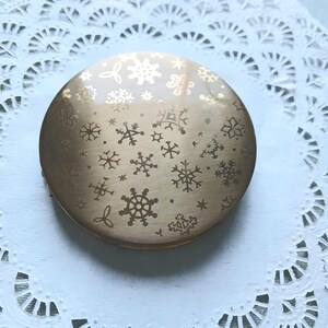 以下が含まれることがあります： 雪の結晶のデザインが施された金色調のコンパクトミラー。ミラーは丸型で、表面は滑らかです。
